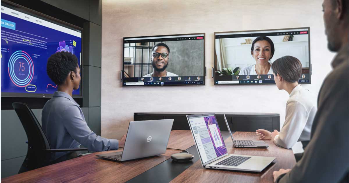 3 people working on Dell laptops at a conference table while 2 monitors on the wall show coworkers