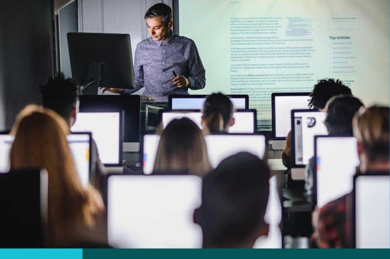 Teacher in front of a screen display presenting to a room full of students on computers