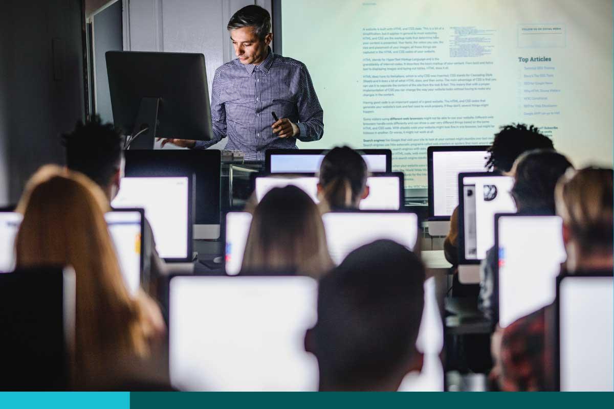 Teacher in front of a screen display presenting to a room full of students on computers